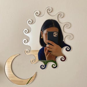 Puede incluir: Un espejo decorativo con un diseño de sol y un acento de media luna. El espejo tiene una superficie clara y reflectante y está montado en una pared blanca. El sol presenta patrones ondulados y arremolinados.
