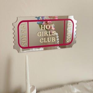 May include: A silver mirror in the shape of a ticket with the words "HOT GIRLS CLUB" in the centre, framed by a pink border. The ticket has the numbers "193405" on each end. A white candle sits in a white decorative holder.
