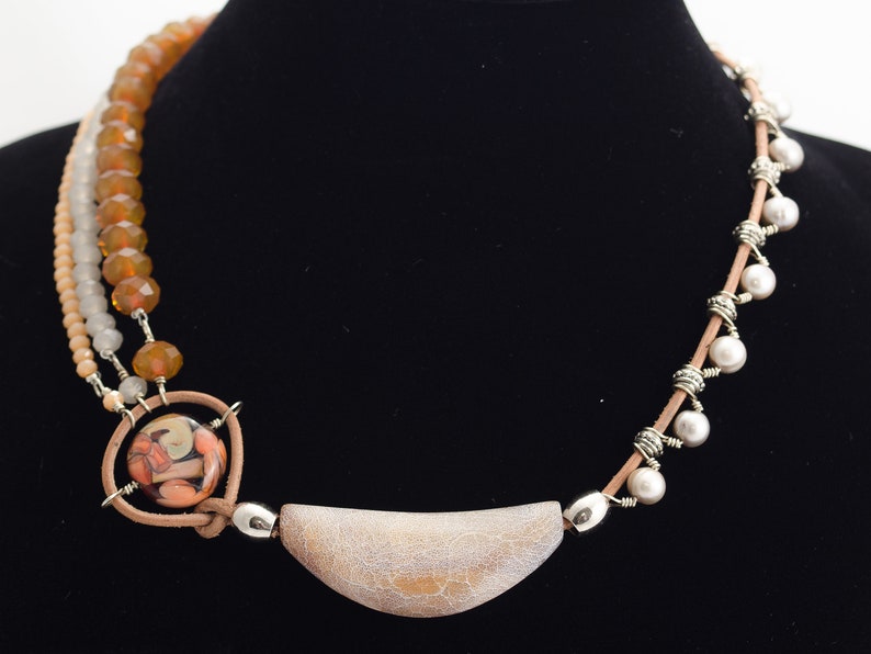 May include: A multi-strand necklace featuring brown, white and cream beads, a large cream-coloured pendant, and a smaller pendant with a colourful glass design.