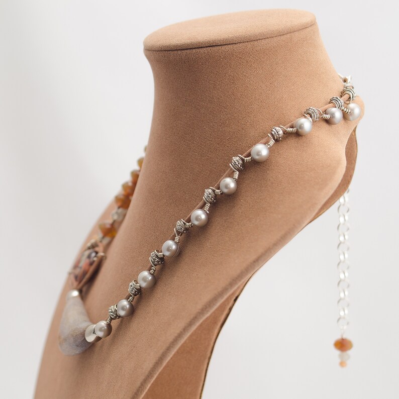 May include: A silver necklace with a long, grey, smooth stone pendant and white pearls. The necklace is on a brown display stand.