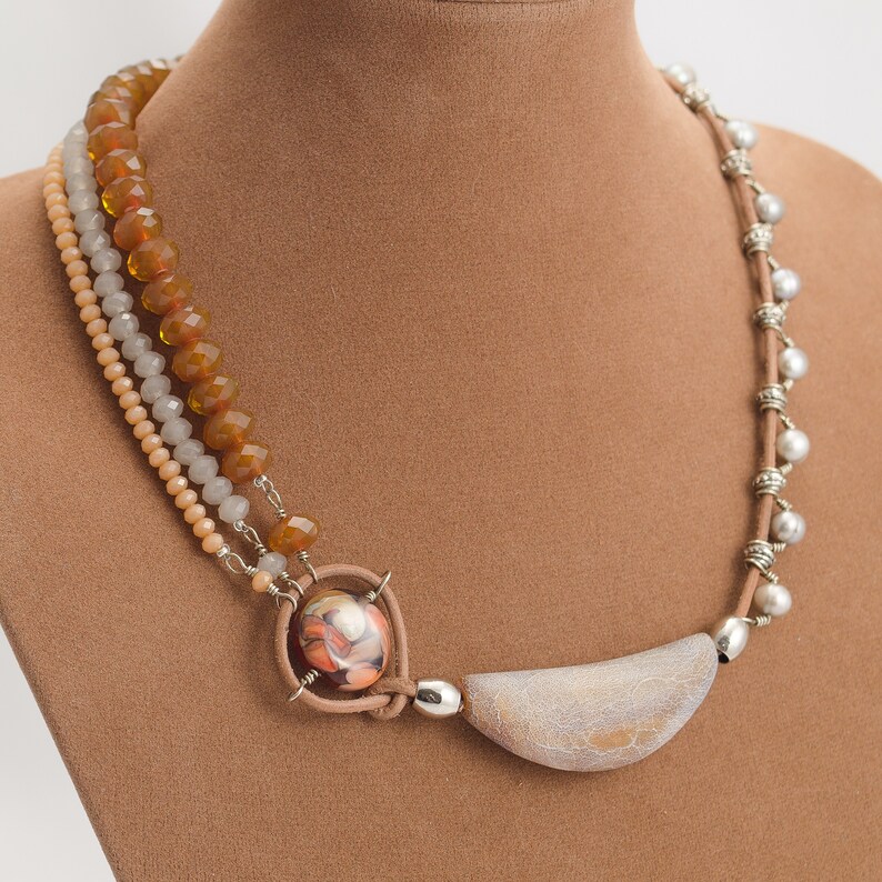 May include: A multi-strand necklace featuring brown, beige, and white beads, a large oval-shaped pendant with a brown, white, and orange design, and a crescent-shaped pendant with a white and grey design.