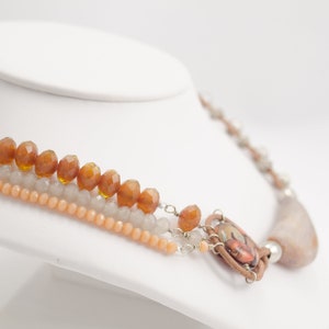 May include: A multi-strand necklace featuring brown, peach, and grey beads. The necklace has a large, brown, oval pendant with a colourful design.