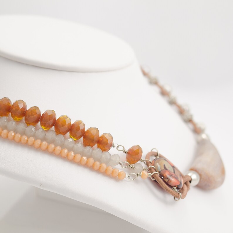 May include: A multi-strand necklace featuring brown, peach, and grey beads. The necklace has a large, brown, oval pendant with a colourful design.