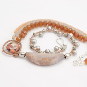 May include: A multi-strand necklace featuring brown leather cord, silver wire, and a variety of beads. The necklace includes a large, light brown agate pendant with a textured surface, a smaller round pendant with a colourful swirl pattern, and several round, light grey beads.