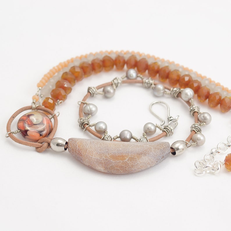 May include: A multi-strand necklace featuring brown leather cord, silver wire, and a variety of beads. The necklace includes a large, light brown agate pendant with a textured surface, a smaller round pendant with a colourful swirl pattern, and several round, light grey beads.