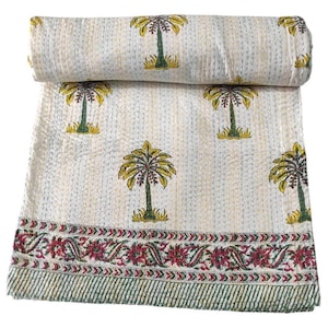 Op de afbeelding: Een opgerolde, crèmekleurige quilt met een herhalend patroon van gele en groene palmbomen. De quilt heeft een decoratieve rand met rode en groene bloemmotieven. De quilt is gemaakt van een zachte, gewatteerde stof.