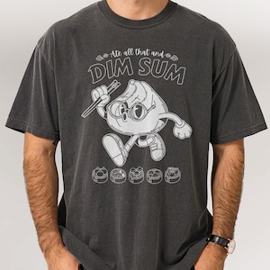 May include: Dark gray t-shirt featuring a cartoon dim sum character holding chopsticks, with the text "Ate all that and DIM SUM." The design includes various dim sum dishes. A casual, food-themed graphic tee.