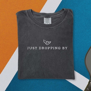 May include: Dark gray t-shirt with the text "JUST DROPPING BY" and a small graphic of a winged creature. The shirt is folded and lies on a blue and orange surface, with a pickleball and paddle visible.