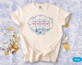May include: Ivory-colored t-shirt with a blue graphic that says "I'm in the mood for" above four heart-suited playing cards. The design includes Asian-inspired imagery. Mahjong tiles are scattered around the shirt on a blue and white patterned background.