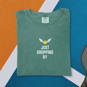 May include: A green t-shirt with the text "JUST DROPPING BY" and a graphic of a pickleball with wings. A yellow pickleball and a gray pickleball paddle are also visible. The shirt is on a blue and orange surface.