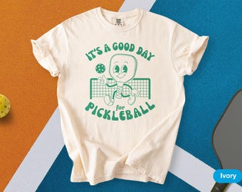 May include: Ivory-colored t-shirt with the text "IT'S A GOOD DAY for PICKLEBALL" in green. The graphic features a cartoon pickleball paddle character. A yellow pickleball and a paddle are also visible.