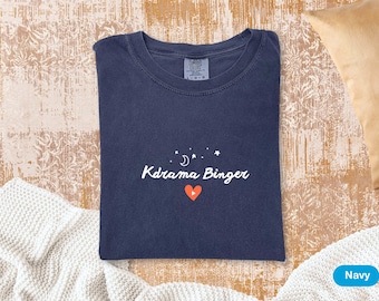 May include: Navy blue t-shirt with the text "Kdrama Binger" in white, along with a crescent moon, stars, and a red heart. The shirt is folded and lies on a textured surface.