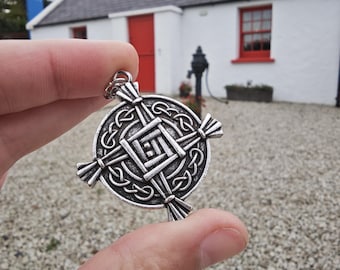 May include: A silver-colored pendant with a Celtic knot design. The pendant is round with a central square knot and four radiating arms. The background shows a white building with a red door and window.