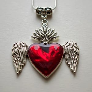 Puede incluir: Collar con colgante plateado con un colgante en forma de corazón con un centro rojo vibrante y alas plateadas a cada lado. El colgante tiene una parte superior plateada decorativa y cuelga de una cadena plateada.
