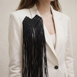 Könnte beinhalten: Ein schwarzer Makramee-Schulterakzent ist an einem weißen Blazer befestigt. Das Makramee besteht aus eng gewebten schwarzen Kordeln, die oben ein herzförmiges Design bilden und über die Schulter fallen. Der Blazer hat einen einzelnen Knopfverschluss.