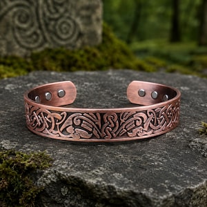 Può includere: Un bracciale a fascia in rame caratterizzato da un intricato motivo a intreccio in rilievo sulla superficie esterna. L'interno del cinturino aperto presenta sei piccoli inserti magnetici circolari di colore scuro vicino alle estremità.
