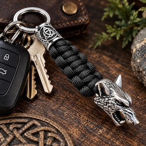 May include: A keychain featuring a black paracord handle with a silver-toned metal wolf head at one end and a cylindrical bead engraved with a triquetra knot symbol at the other, attached to a circular metal ring holding several keys.
