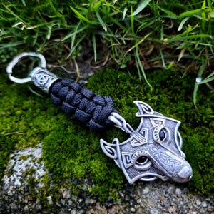 May include: A silver-colored wolf head keychain with black accents and a braided black cord. The keychain is lying on a bed of green moss and grass. The wolf head has intricate Celtic knot designs.