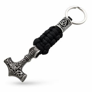 Könnte beinhalten: Ein silberfarbener Metall-Thorhammer-Anhänger-Schlüsselanhänger mit aufwendigen keltischen Knotenmustern. Der Hammer ist an einer schwarzen geflochtenen Kordel und einem silbernen Schlüsselring befestigt. Das Design ist detailliert und im Wikinger-Stil gehalten.