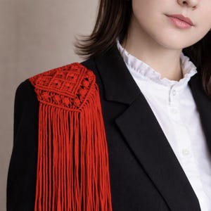 Op de afbeelding: Een zwarte blazer met een rode macramé schouderdetail. Het schouderstuk heeft een geometrisch ontwerp en lange franjes. De blazer wordt gedragen over een wit overhemd met kraag, wat een contrast in texturen en kleuren creëert.