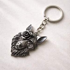 Pode incluir: Um porta-chaves de metal prateado com um pendente detalhado e esculpido de uma cabeça de lobo. O lobo tem uma flor de rosa posicionada entre as orelhas. O pendente está preso a um anel circular padrão por uma corrente curta de elos.