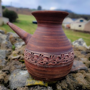 May include: A brown ceramic pitcher with a long spout and a decorative floral band. The pitcher has a textured surface. It is set against a natural, outdoor backdrop.