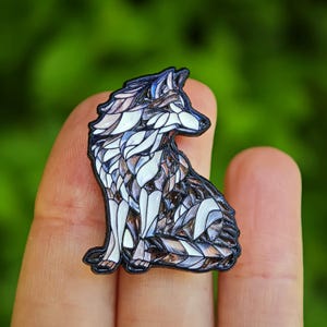 Op de afbeelding: Een gedetailleerde emaille pin met een zittende wolf. De pin heeft een zwarte omtrek en is gevuld met een mozaïekachtig patroon van witte, bruine en blauwe vormen. De wolf kijkt naar rechts tegen een vage groene achtergrond.