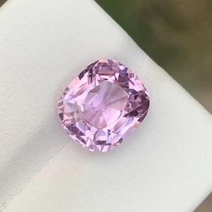 May include: A faceted, light pink gemstone with a cushion cut. The gem is set against a white surface, with a blurred green background. The gem's facets reflect light, creating a sparkling effect. This gemstone is likely for jewelry.