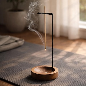 以下が含まれることがあります： 木製の香立てと黒い金属製のスタンド。 燃えているお香のスティックが立っています。 お香から煙が立ち上り、穏やかな雰囲気を作り出しています。 ホルダーは灰色のマットの上に置かれています。