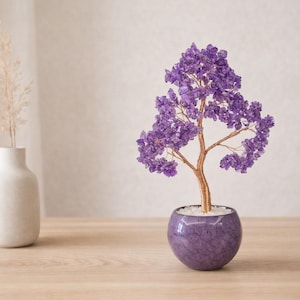 May include: A decorative amethyst crystal tree with a copper wire trunk and branches, set in a round, purple, speckled pot. A white vase with dried grass sits to the left on a wooden table. The tree is a unique home decor item.