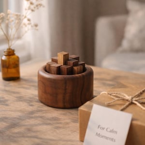 Może przedstawiać: Drewniana układanka z cylindryczną podstawą i małymi, kwadratowymi klockami. Klocki tworzą trójwymiarową strukturę. Mała, bursztynowa szklana butelka z suszonymi kwiatami i pudełko prezentowe z napisem "For Calm Moments" są również obecne.