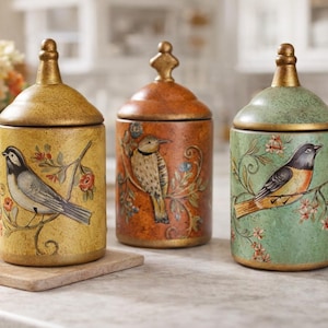 Pode incluir: Três potes decorativos de cerâmica com tampas, cada um com uma ilustração de pássaro diferente. Os potes são pintados em tons de amarelo, laranja e verde, com detalhes dourados. São exibidos em uma bancada, para decoração de cozinha.