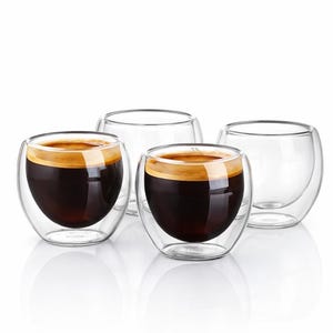 Może przedstawiać: Cztery przezroczyste, dwuścienne szklanki do espresso. Dwie szklanki są wypełnione ciemną kawą i zwieńczone warstwą cremy. Pozostałe dwie szklanki są puste. Szklanki mają zaokrąglony kształt.