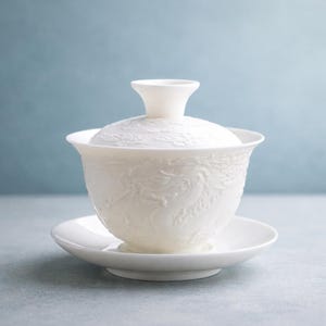 May include: White porcelain gaiwan tea set with a matching lid and saucer. The gaiwan features a detailed embossed dragon design. The set is designed for tea ceremonies and is placed on a light gray surface against a blue background.
