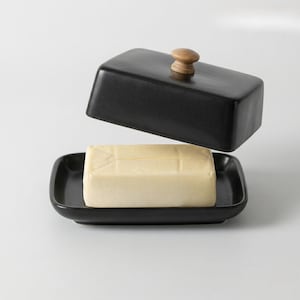 May include: A black ceramic butter dish with a wooden knob on the lid. The dish holds a rectangular block of butter. The lid is slightly raised, revealing the butter. The butter dish is on a white background.