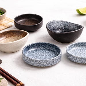 Puede incluir: Una colección de pequeños cuencos de cerámica en varios colores y patrones. Los cuencos son en tonos de negro, marrón y azul y blanco moteado. También se ven palillos y sushi.