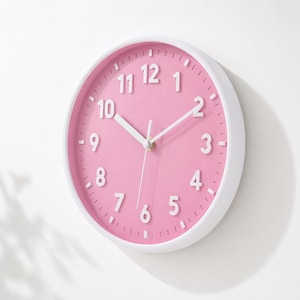 May include: A round wall clock with a white frame and a pink face. The clock features white numerals and hands, with a second hand. The clock is a decorative item for a home or office.