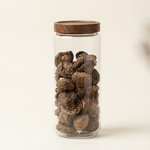 Könnte beinhalten: Ein klares Glas mit getrockneten Shiitake-Pilzen. Das Glas hat einen Holzdeckel und steht vor einem neutralen Hintergrund. Die Pilze haben eine Mischung aus braunen und beigen Farben.