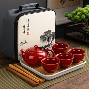 May include: A red ceramic tea set with a teapot, four cups, and a serving tray. The set includes a carrying case with a landscape painting. Two bamboo sticks and a brown cloth are also included.