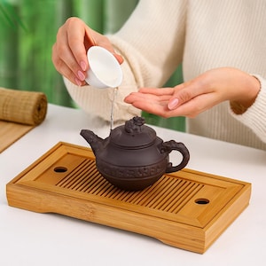May include: A brown ceramic teapot with a decorative floral design sits on a bamboo tea tray. A small white cup pours liquid into the teapot. The tray has a rectangular shape with drainage slots and a round hole.