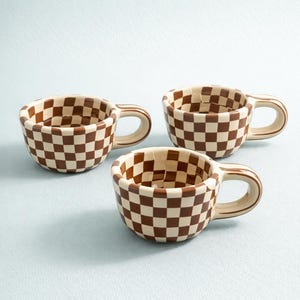 Puede incluir: Tres tazas de espresso de cerámica con un patrón de tablero de ajedrez marrón y crema. Cada taza tiene un asa redondeada y un diseño de tablero de ajedrez a juego en el interior. Las tazas se muestran sobre una superficie azul claro.