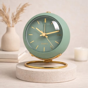 Könnte beinhalten: Eine hellgrüne Quarzuhr mit goldfarbenen Zeigern und Akzenten. Die Uhr hat ein rundes Zifferblatt und steht auf einem goldfarbenen Metallständer. Das Wort "QUARTZ" ist auf dem Zifferblatt aufgedruckt. Die Uhr steht auf einem kleinen, runden, hellfarbenen Steinsockel.