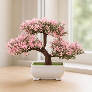 May include: A small bonsai tree with pink flowers and green leaves. The tree has a brown trunk and branches, and is planted in a white rectangular pot. The pot is on a wooden surface.