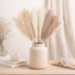 Könnte beinhalten: Eine beige strukturierte Vase enthält künstliches Pampasgras in Creme-, Beige- und Brauntönen. Die Vase steht auf einer Holzoberfläche, daneben eine kleine weiße Keramikschale und Bücher. Eine Kette aus Holzperlen ist ebenfalls zu sehen.