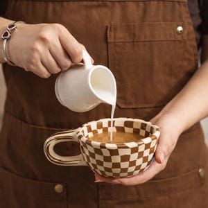 Könnte beinhalten: Ein weißer Sahneaufgießer gießt Milch in eine braun-creme karierte Kaffeetasse. Die Tasse hat einen passenden karierten Griff. Die Person, die die Tasse hält, trägt eine braune Schürze.