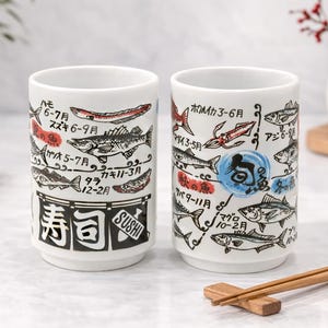 Puede incluir: Dos tazas de sushi de cerámica blanca con ilustraciones en negro y rojo de varios peces y texto japonés. La palabra "SUSHI" está impresa en negro. Palillos chinos descansan sobre un soporte de madera.