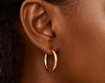 May include: Close-up of a gold hoop earring. The earring is a simple, polished design with a rounded shape. The earring is worn on a person's ear, and the gold color provides a bright contrast to the skin tone.