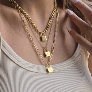 May include: A layered gold necklace set featuring three chains. Each chain has a different design, including a curb chain and a paperclip chain. Each chain has a small padlock pendant with an engraved initial. The necklaces are worn on a person.