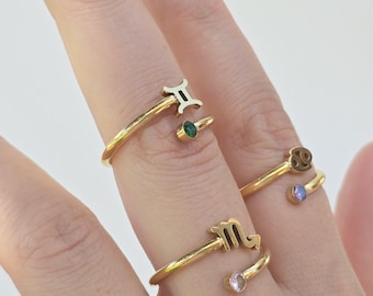 May include: Gold-toned adjustable rings featuring zodiac symbols: Gemini, Cancer, and Scorpio. Each ring has a small gemstone accent in green, purple, and pink. The rings are displayed on a hand against a white background.