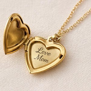 Könnte beinhalten: Eine goldfarbene herzförmige Medaillon-Halskette, geöffnet, um die eingravierten Worte "Love Mom" zu zeigen. Das Medaillon hängt an einer zarten Kette. Ein klassisches Schmuckstück.